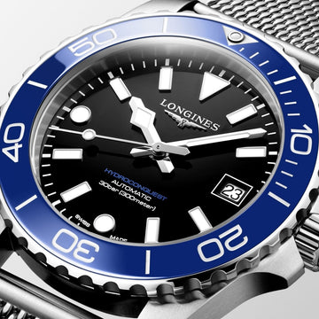 Close-up of a Longines HydroConquest watch showing black face, luminous hands, blue rotating bezel, and steel mesh strap